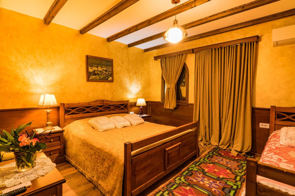 A bedroom with a wooden bed, a nightstand with a lamp, a bedside table with a vase of flowers, and a window with curtains.