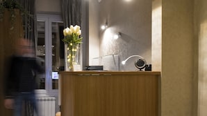 A reception desk with a vase of white flowers, a wall-mounted lamp, and a digital screen. - Relais Bocca di Leone (Rome)
