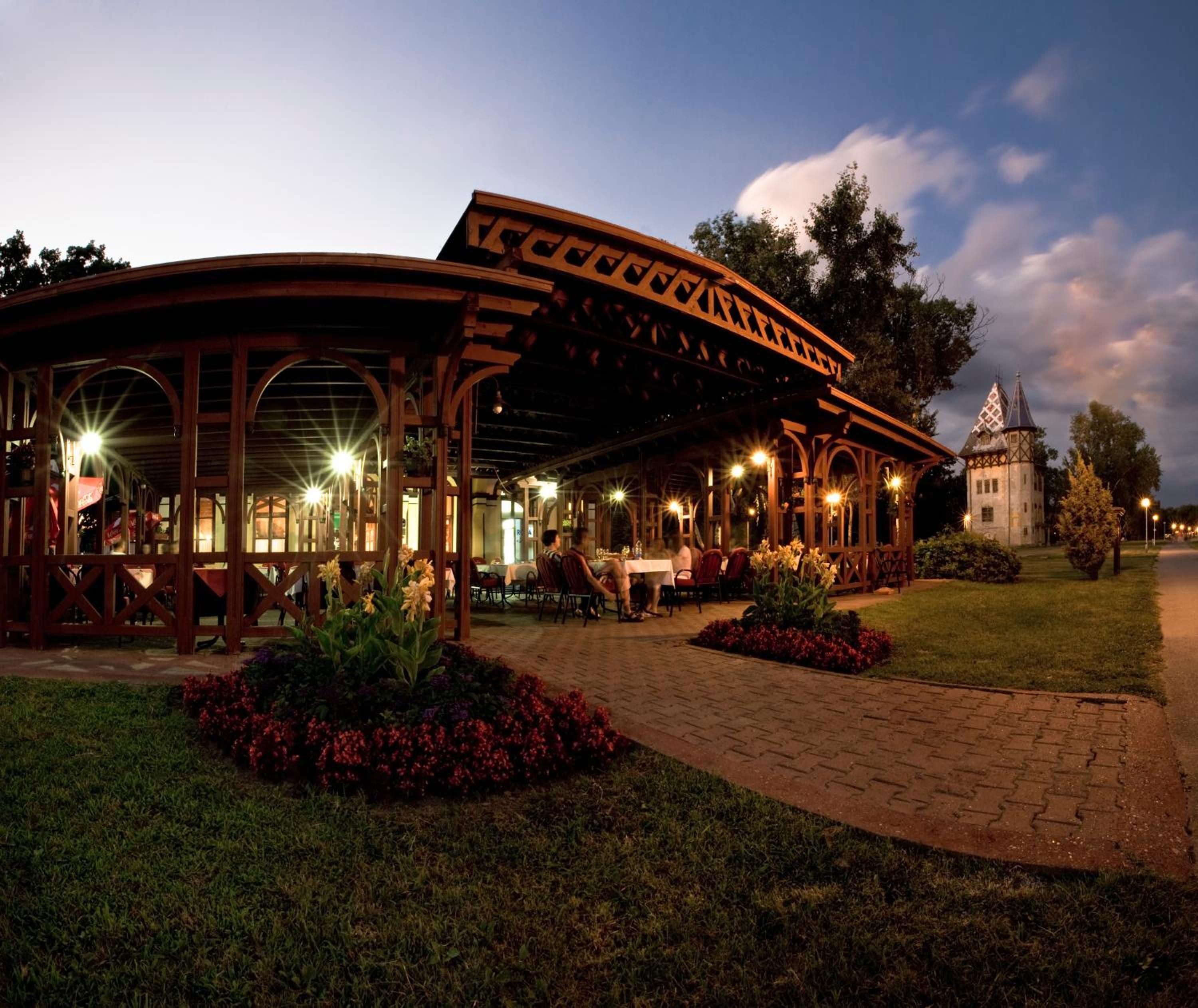 A covered outdoor seating area with tables and chairs, surrounded by flower beds and a paved walkway.