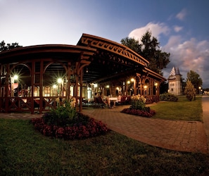 A covered outdoor seating area with tables and chairs, surrounded by flower beds and a paved walkway. - Hotel Park (Palic)