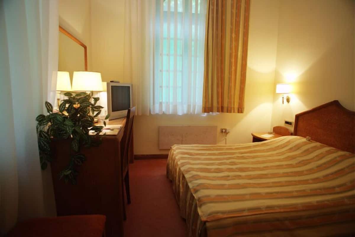 Quarto de hotel com uma cama, uma escrivaninha com computador, uma cadeira, planta em vaso, janela com cortinas e mesa de cabeceira com abajur.