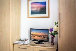 A room with a wooden desk, a monitor displaying a marina scene, a framed picture above the desk, and a vase with flowers to the right.