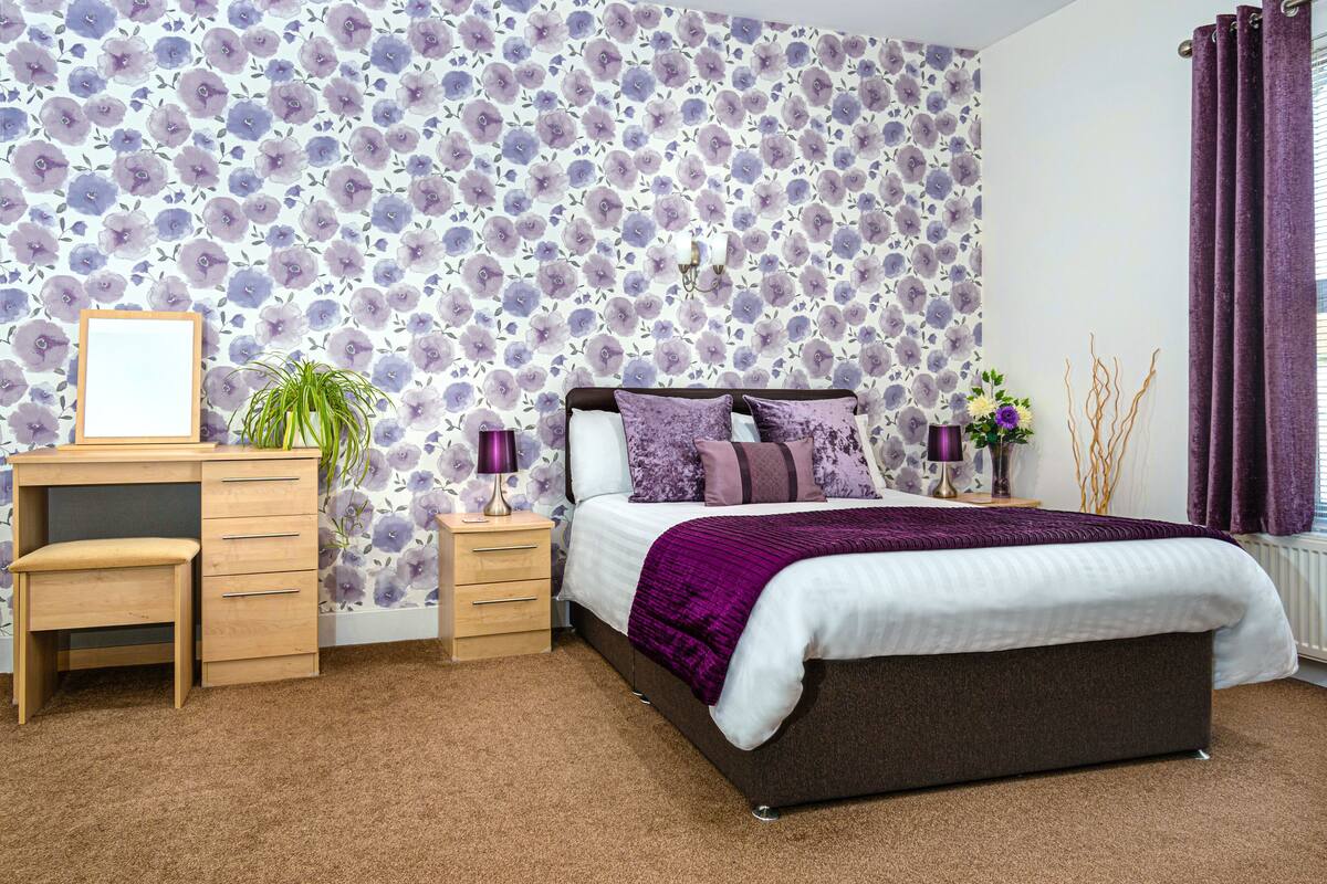 A bedroom with a floral wallpaper, a bed with a purple blanket, a desk with a chair, and a nightstand with a lamp.
