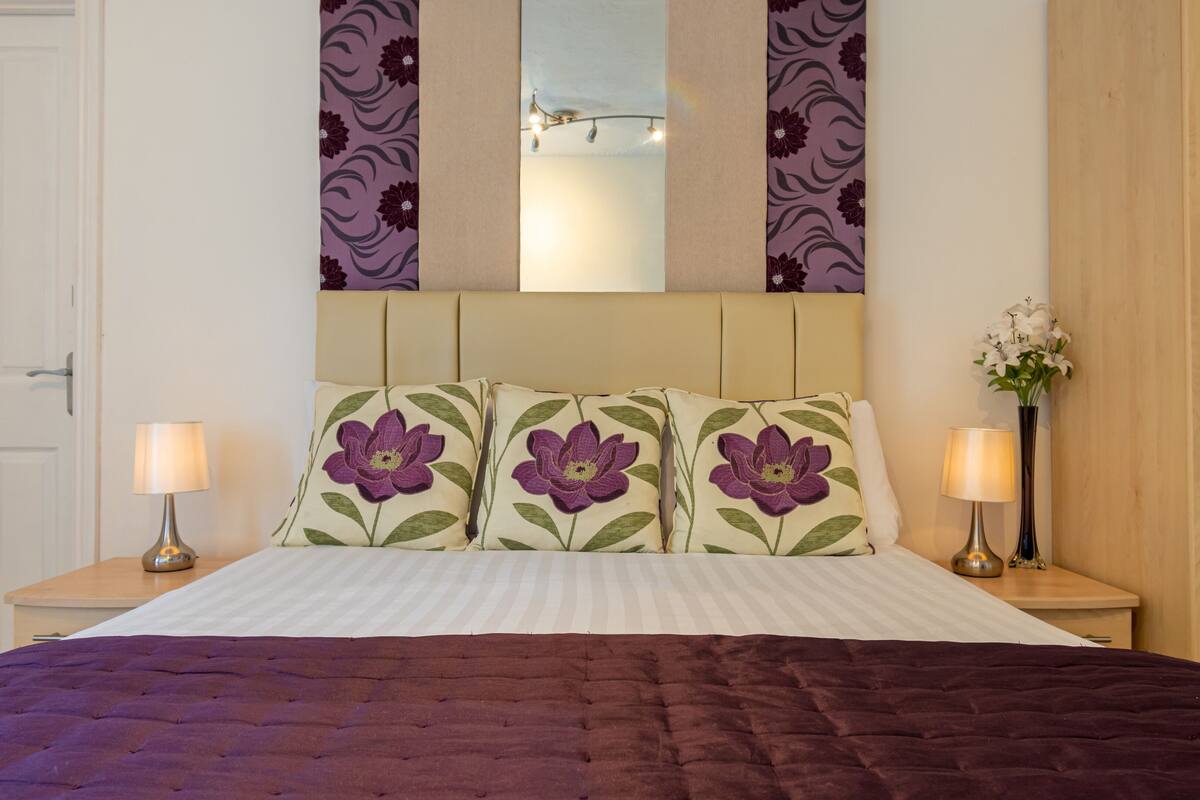 A neatly made bed with floral pillows, a bedside lamp, and a vase with flowers on the nightstand.