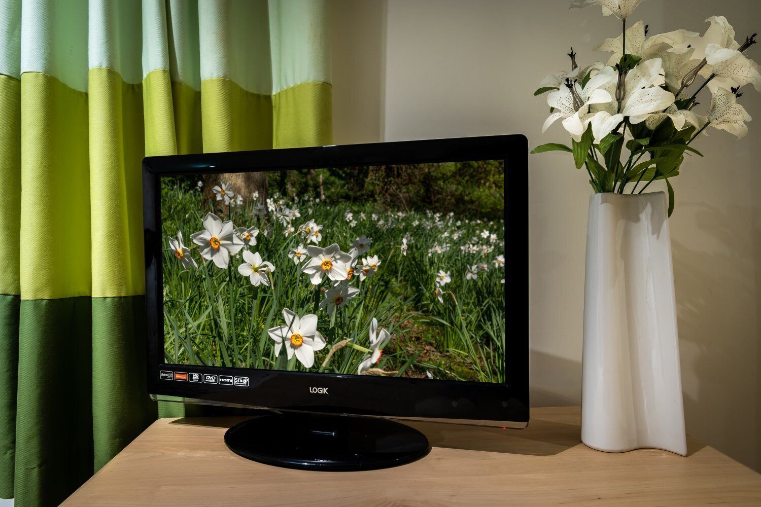 A television on a wooden stand, a white vase with flowers, and green curtains.