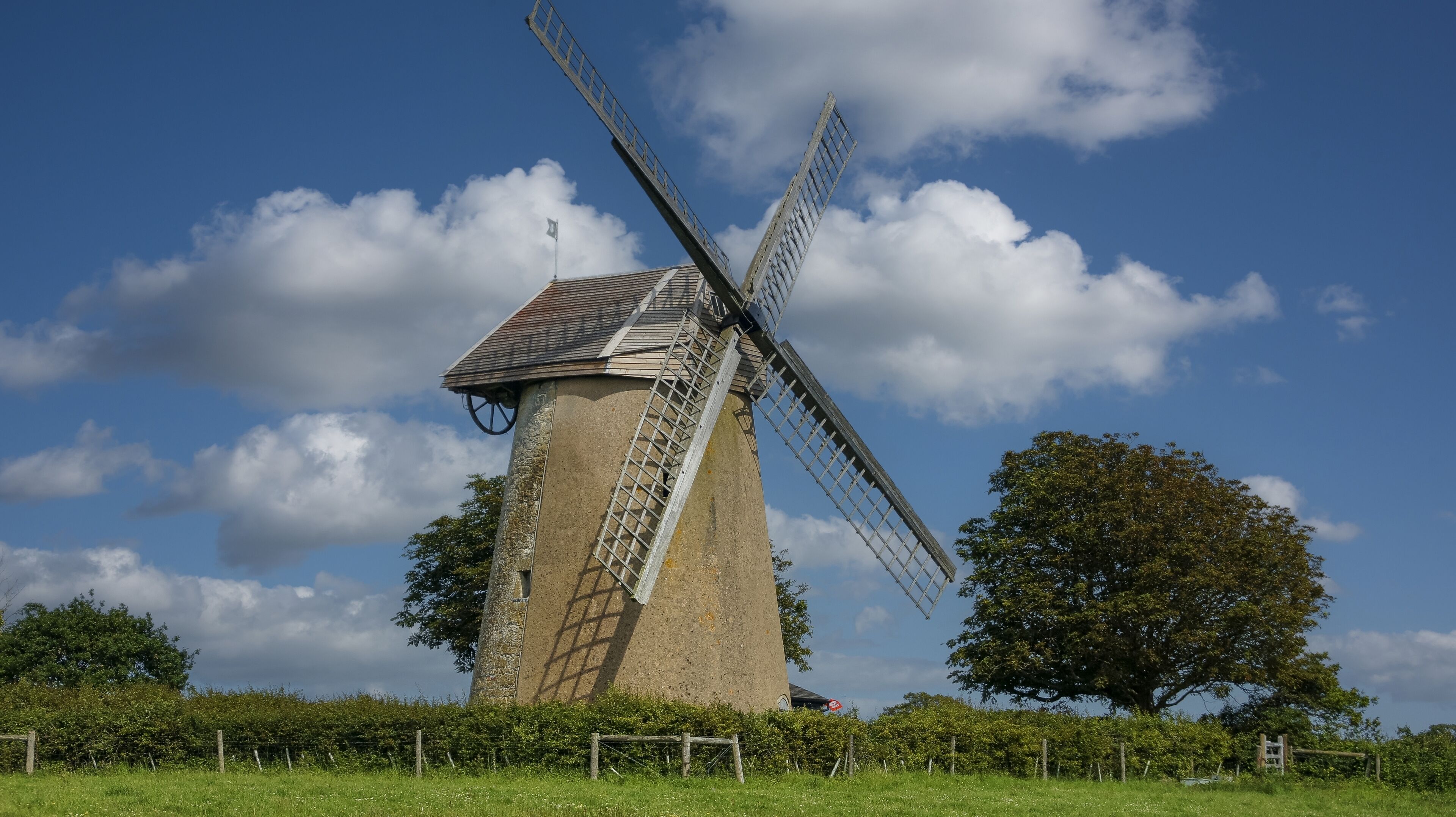A traditional windmill with a metal lattice structure, set against a clear blue sky with scattered clouds.