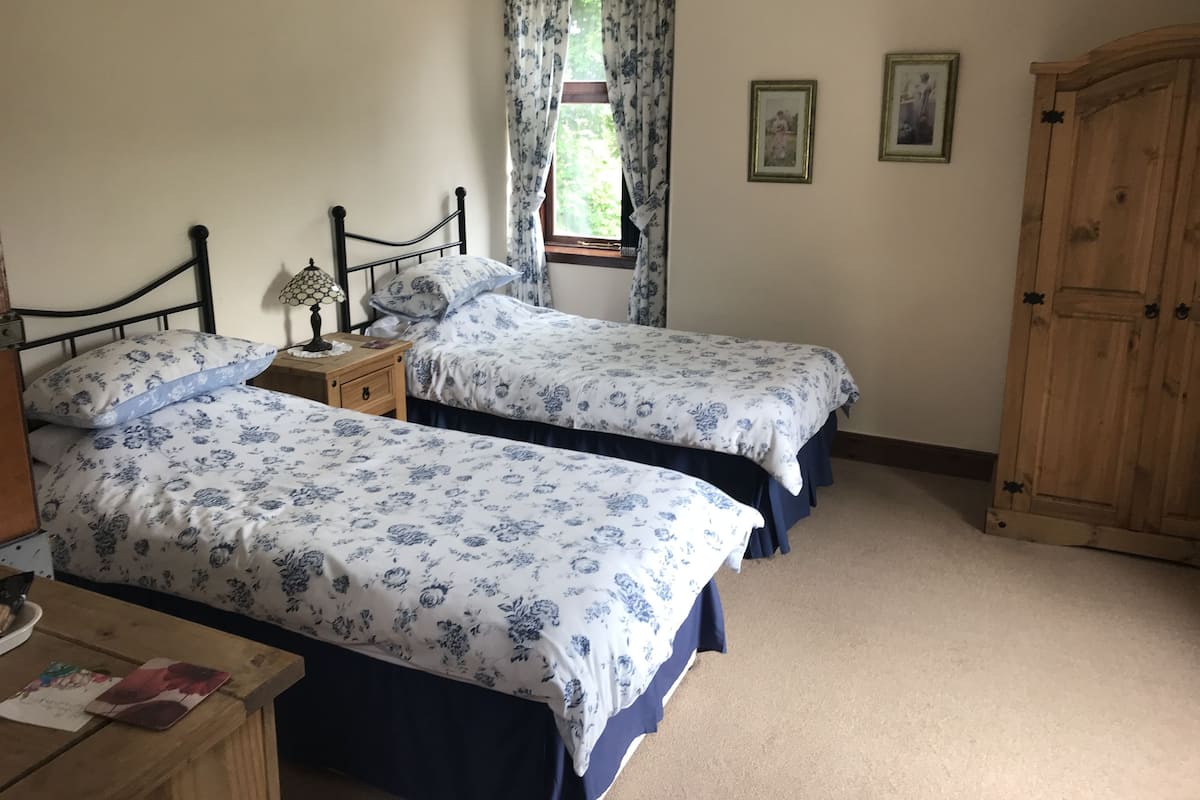 A bedroom with two single beds, a wooden wardrobe, a nightstand with a lamp, and a window with curtains.
