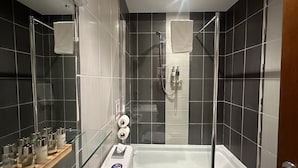 A modern bathroom with a glass-enclosed shower, a white bathtub, and a glass shelf with toiletries. - The Bell Guest House (Peterlee)