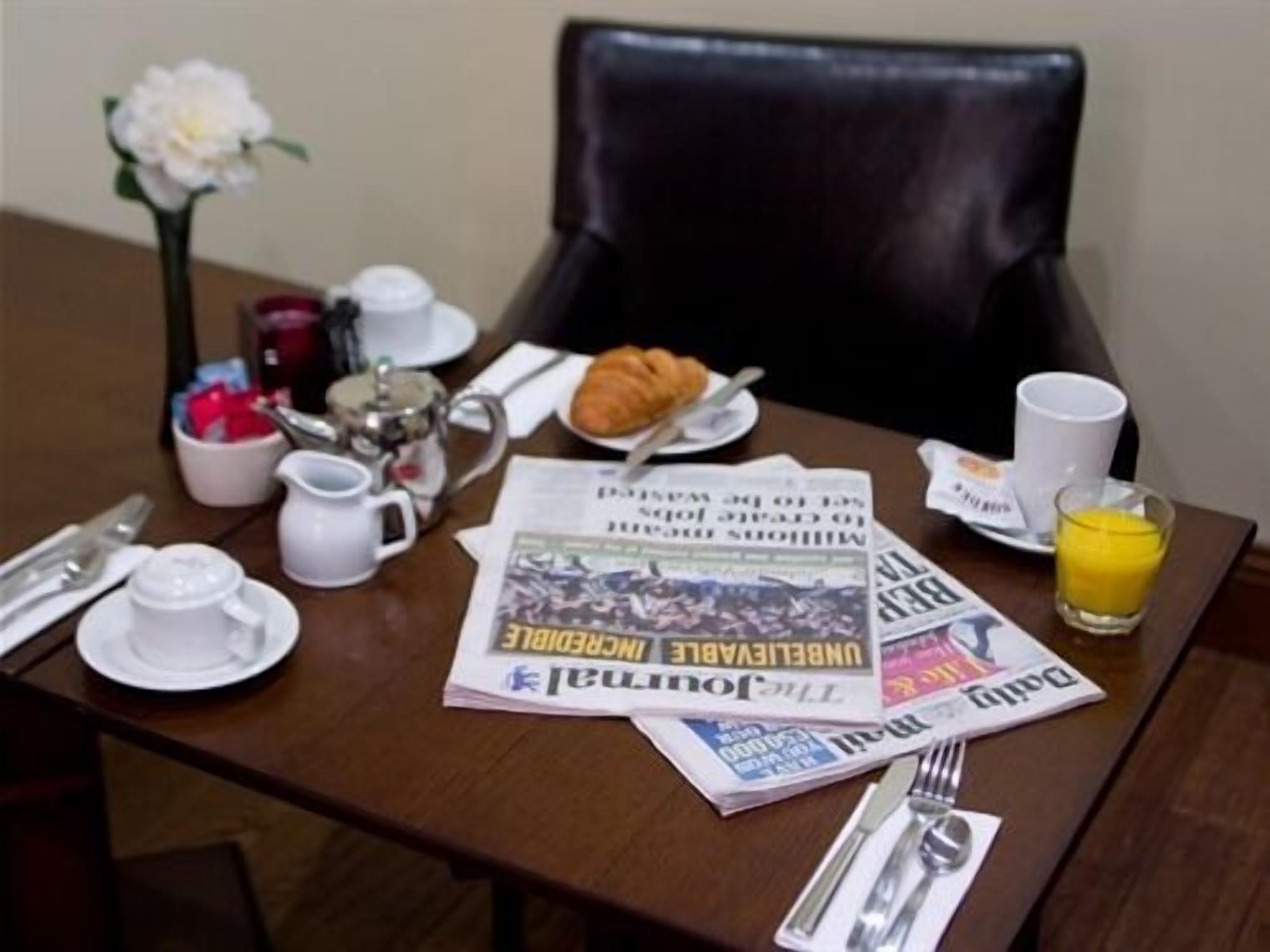 A breakfast table with a croissant, coffee, juice, newspapers, and a vase with a flower.