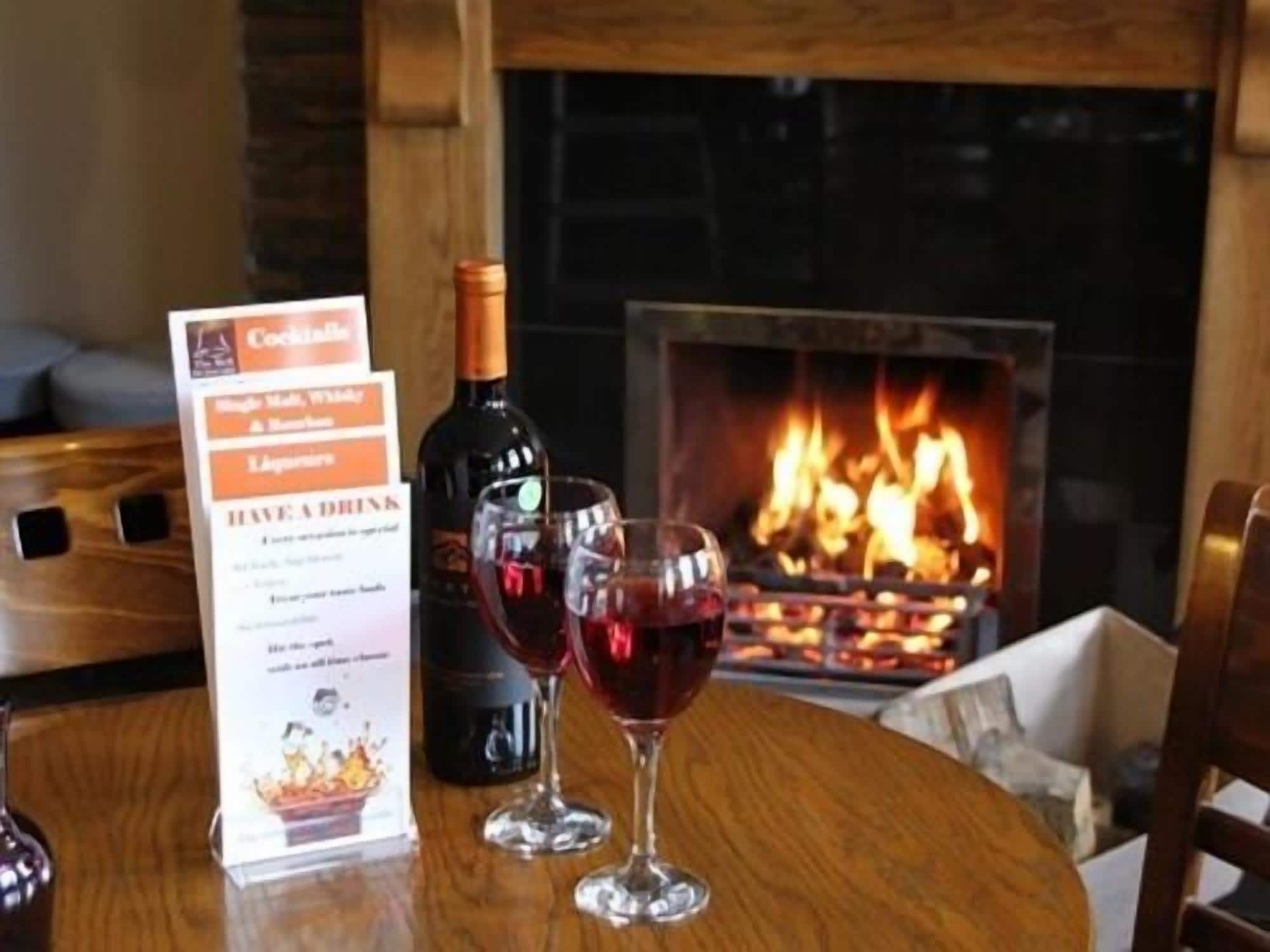 Un tablero de menú con opciones de cócteles, una botella de vino y dos copas de vino tinto sobre una mesa de madera con una chimenea al fondo.