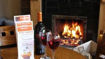 Un tablero de menú con opciones de cócteles, una botella de vino y dos copas de vino tinto sobre una mesa de madera con una chimenea al fondo.
