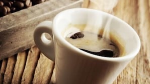 A white cup of coffee with a spoon, placed on a wooden surface with a coffee bean container in the background. - The Bell Guest House (Peterlee)
