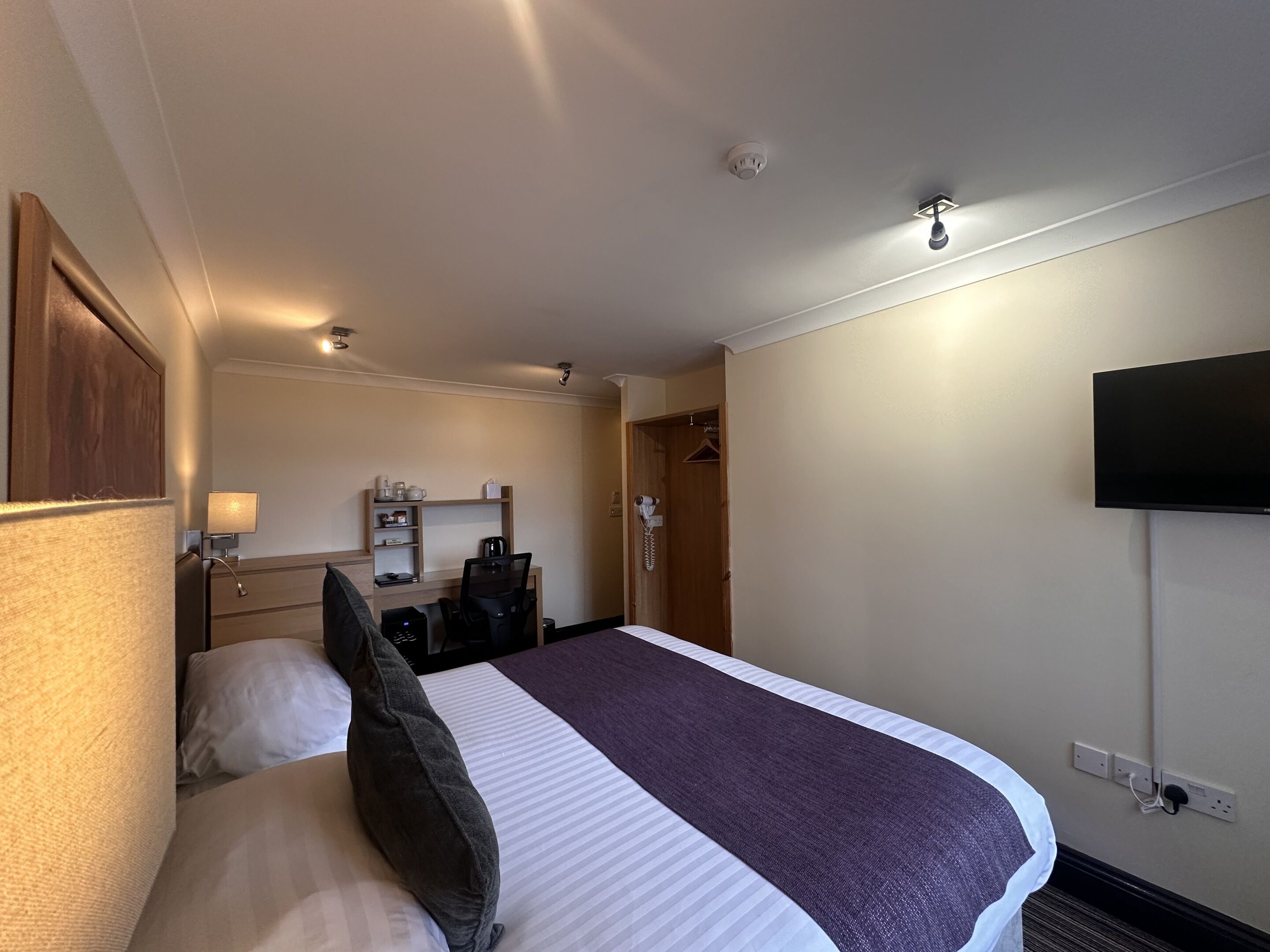 Habitación de hotel con cama, mesita de noche, lámpara y televisor montado en la pared.
