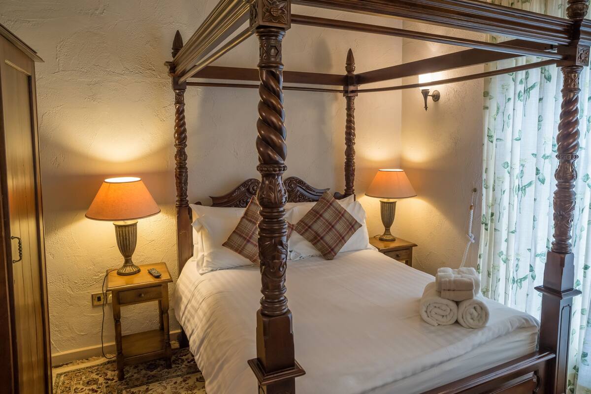 A four-poster bed with two bedside tables, each with a lamp, and a window with floral curtains.