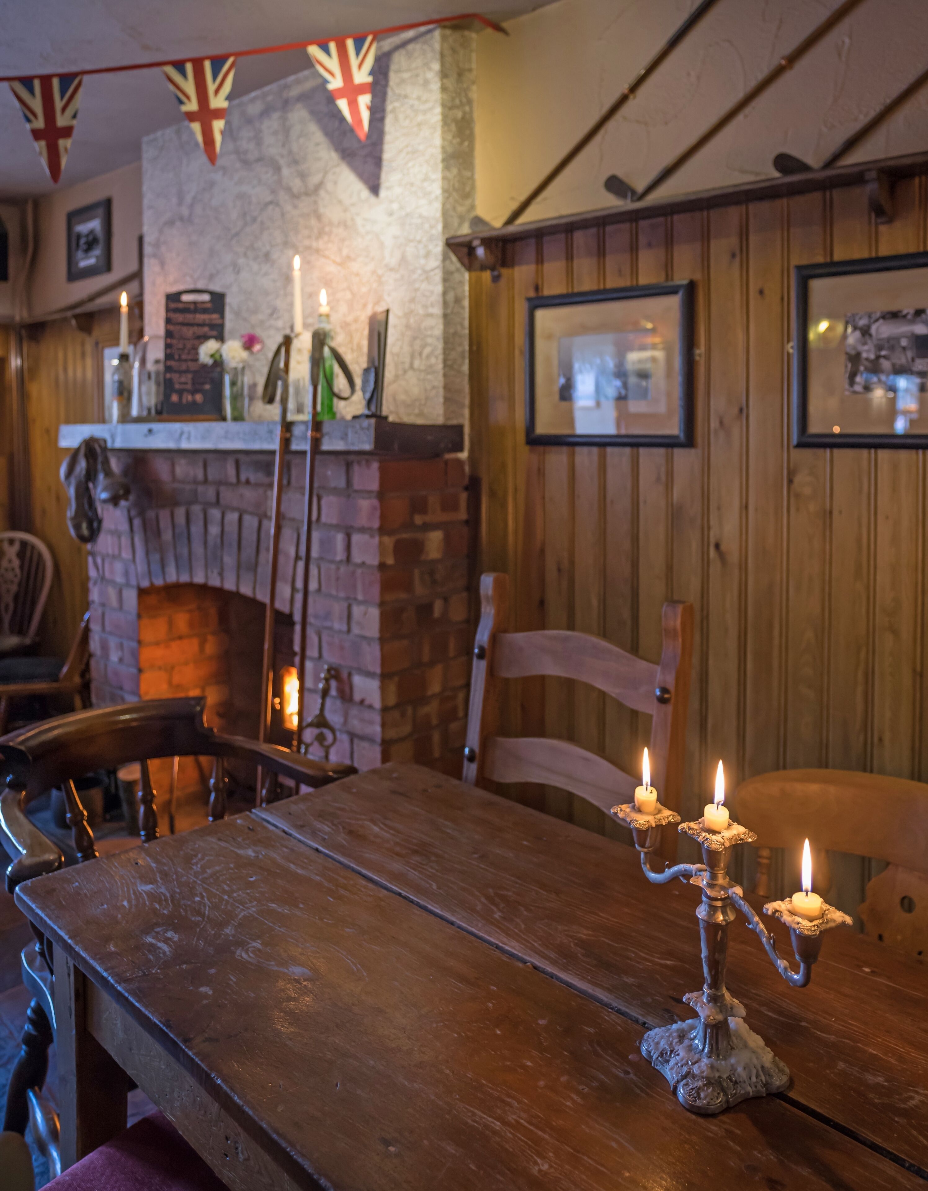 Interior de madera con chimenea de ladrillo, sillas de madera, una mesa con velas y una pared con cuadros.
