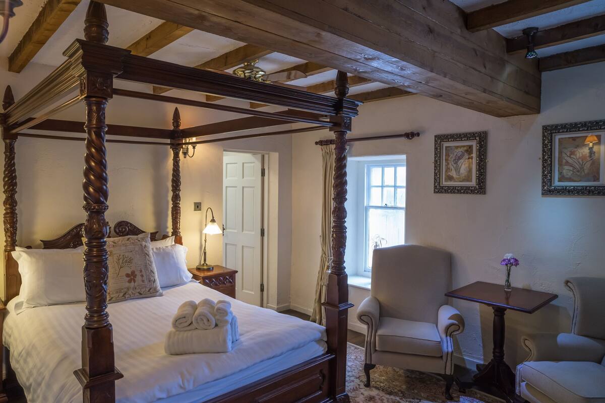 A four-poster bed with a white bedspread and pillows, a wooden nightstand with a lamp, a chair, and a small table with a vase of flowers.