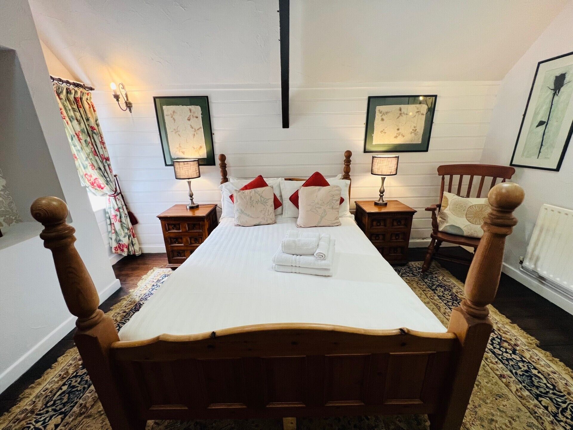A neatly made bed with white linens and red pillows, flanked by wooden nightstands with lamps, in a room with patterned carpet and framed artwork on the walls.