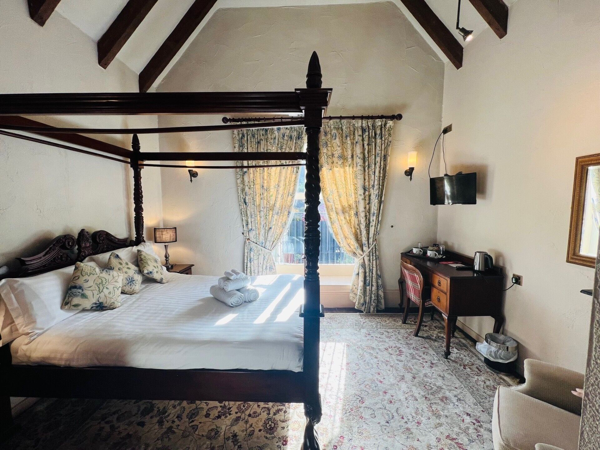 A four-poster bed with a canopy, a desk with a chair, a television, and a window with curtains.