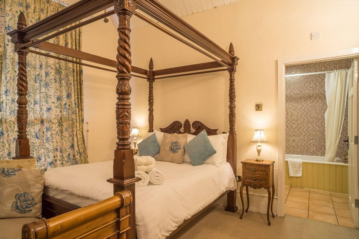 A four-poster bed with a wooden headboard, a bedside table with a lamp, and a bathroom with a tiled shower.