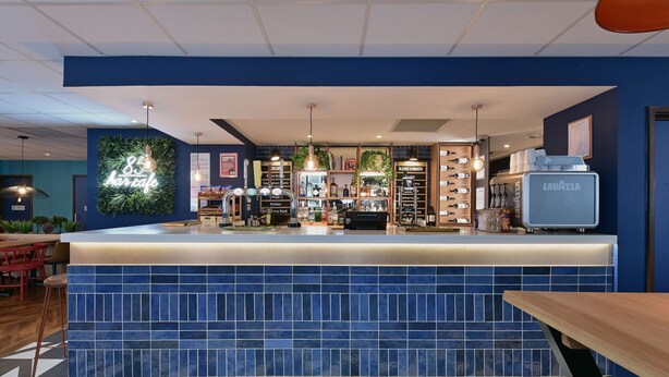 A modern bar area with a blue-tiled counter, a neon sign, and a variety of bottles and glasses on shelves. - Travelodge Cardiff Central Queen Street (Cardiff)