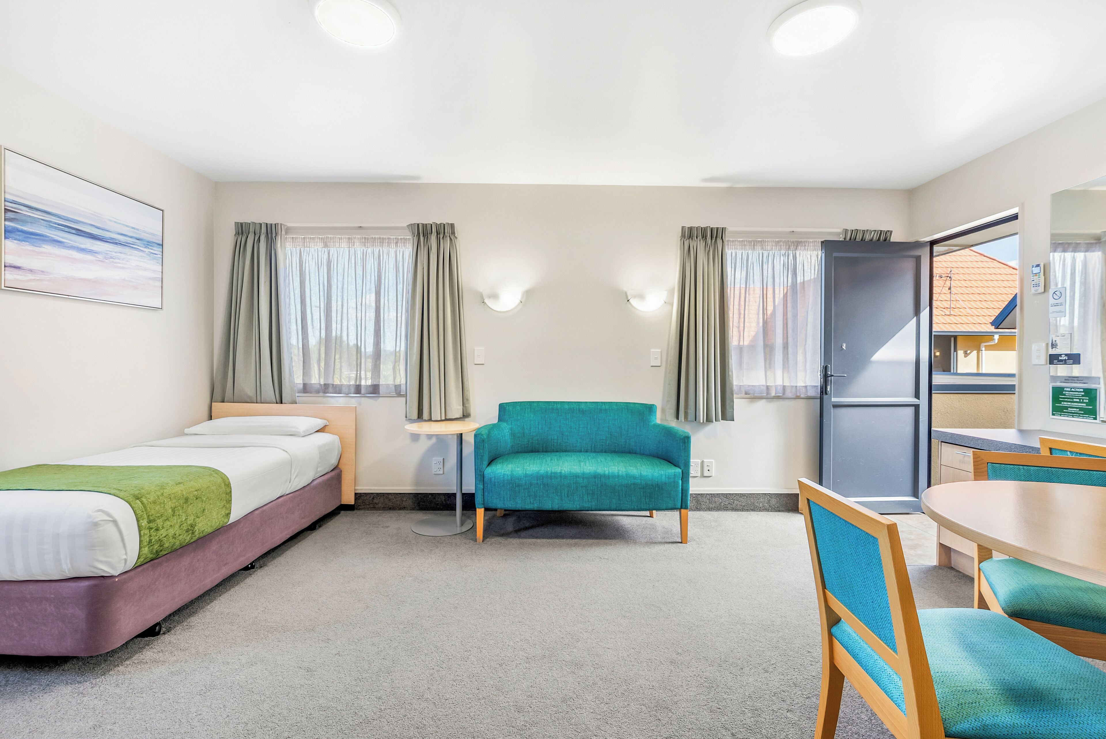 A hotel room with a bed, a teal armchair, a small table, and a window with curtains.