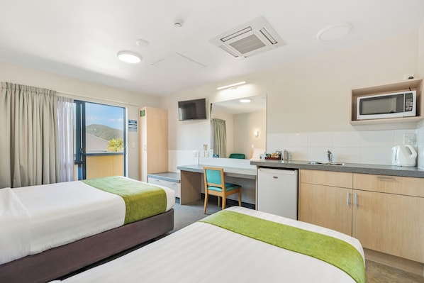 A hotel room with two beds, a desk, a chair, a microwave, a sink, and a view of the outdoors.