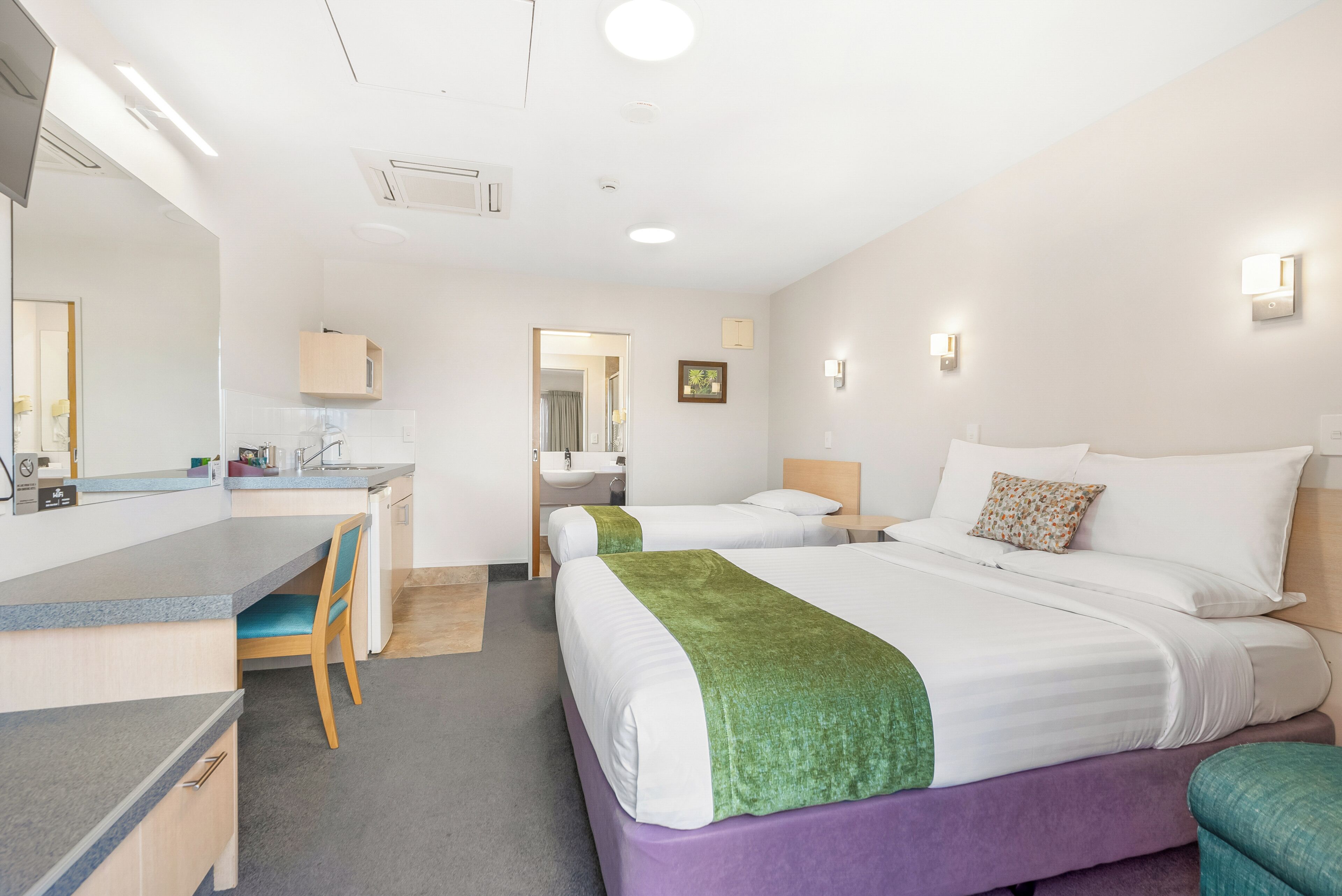 A hotel room with a desk, two beds, a bathroom, and a hallway.