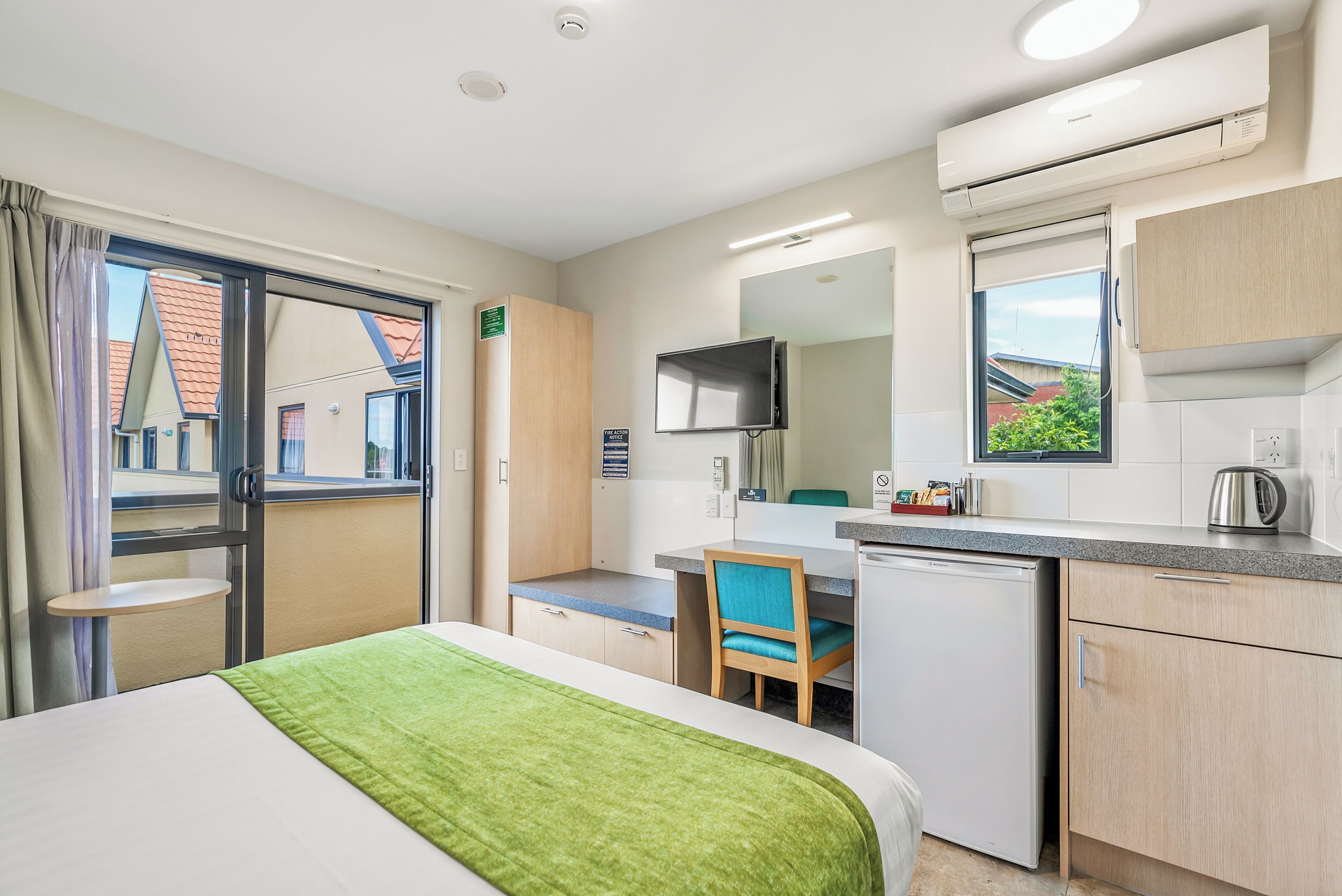 Habitación de hotel con cama, escritorio, silla, televisor, kitchenette y ventana con vistas a edificios.