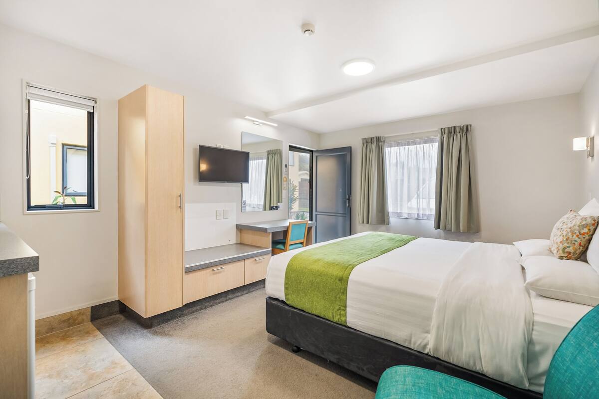 Una habitación de hotel con cama, escritorio, televisor y ventana con cortinas.