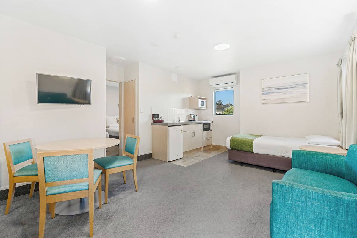 Habitación de hotel compacta con zona de comedor, kitchenette y cama con una manta verde.