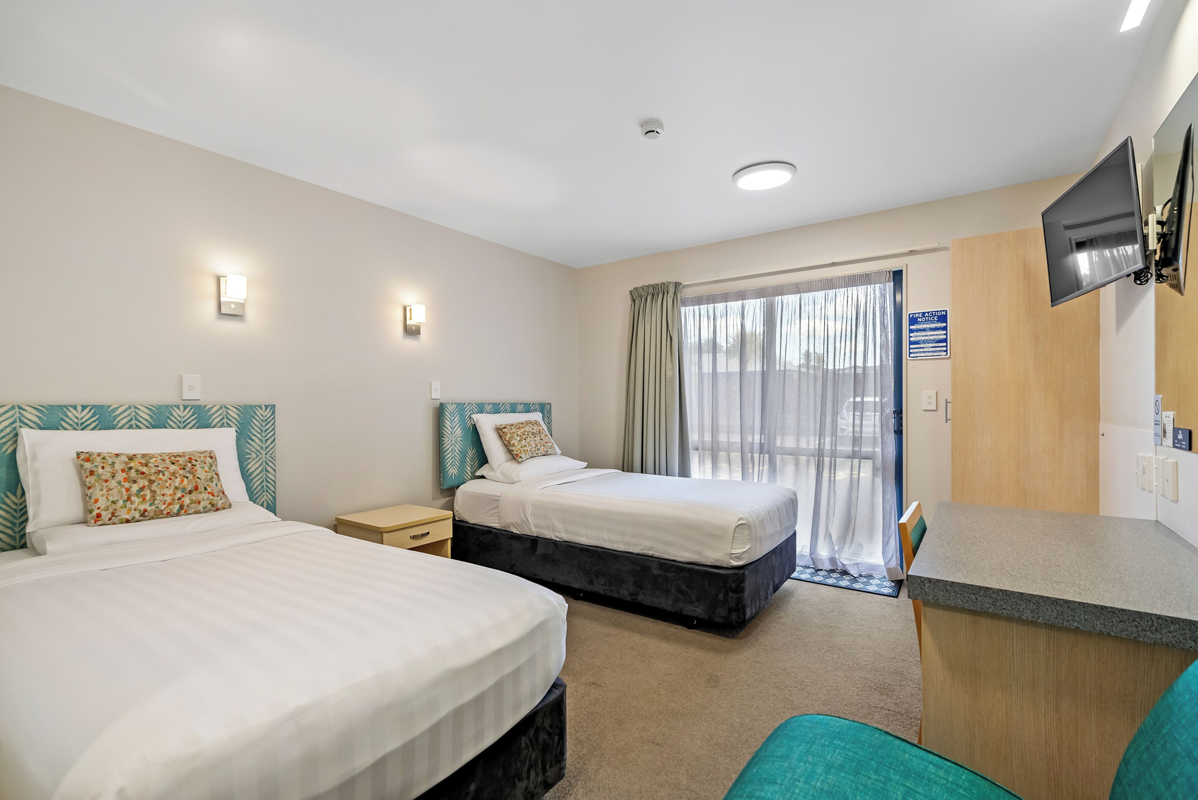 A hotel room with two beds, a desk, a TV, and a sliding door to a balcony.