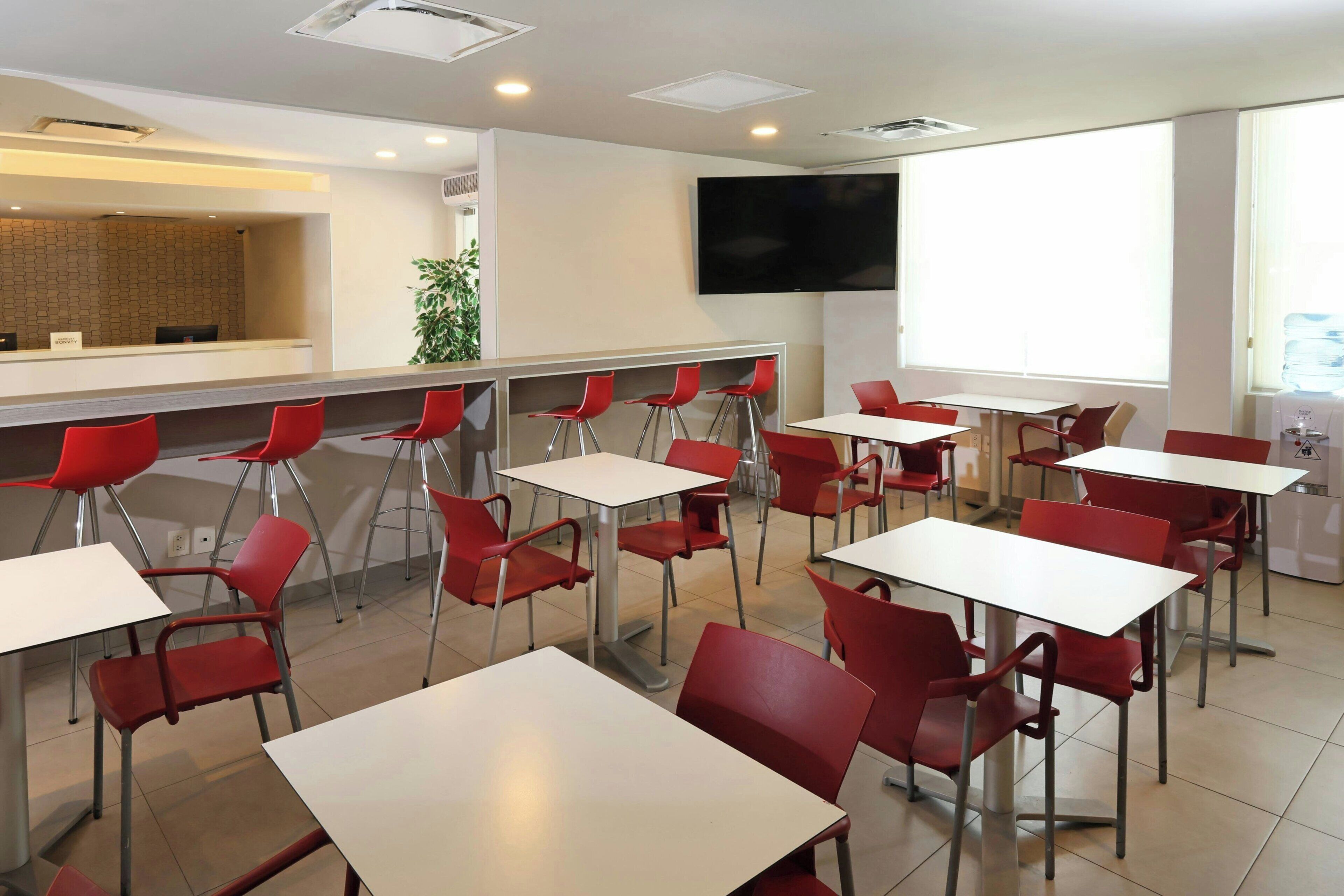 A modern dining area with red chairs and white tables, a reception desk, and a large TV screen.