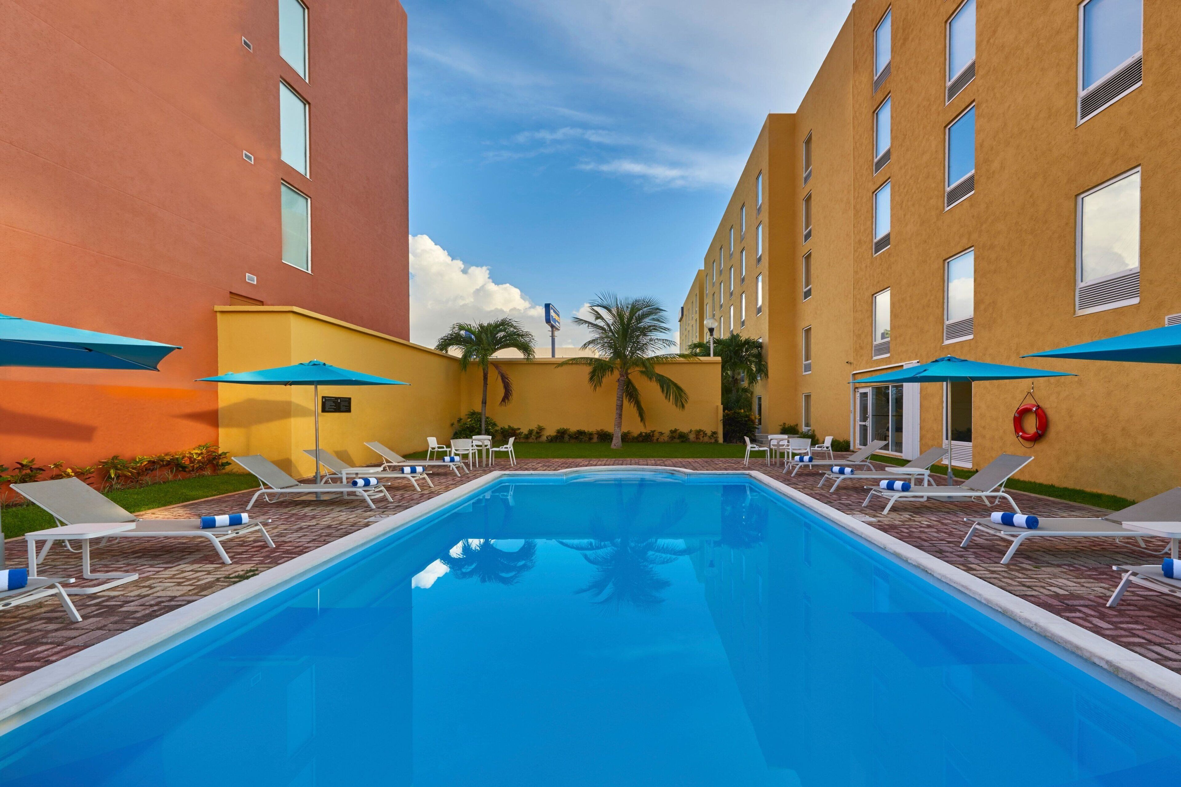 Piscina com espreguiçadeiras e guarda-sóis, cercada por edifícios, um caminho de tijolos e palmeiras.