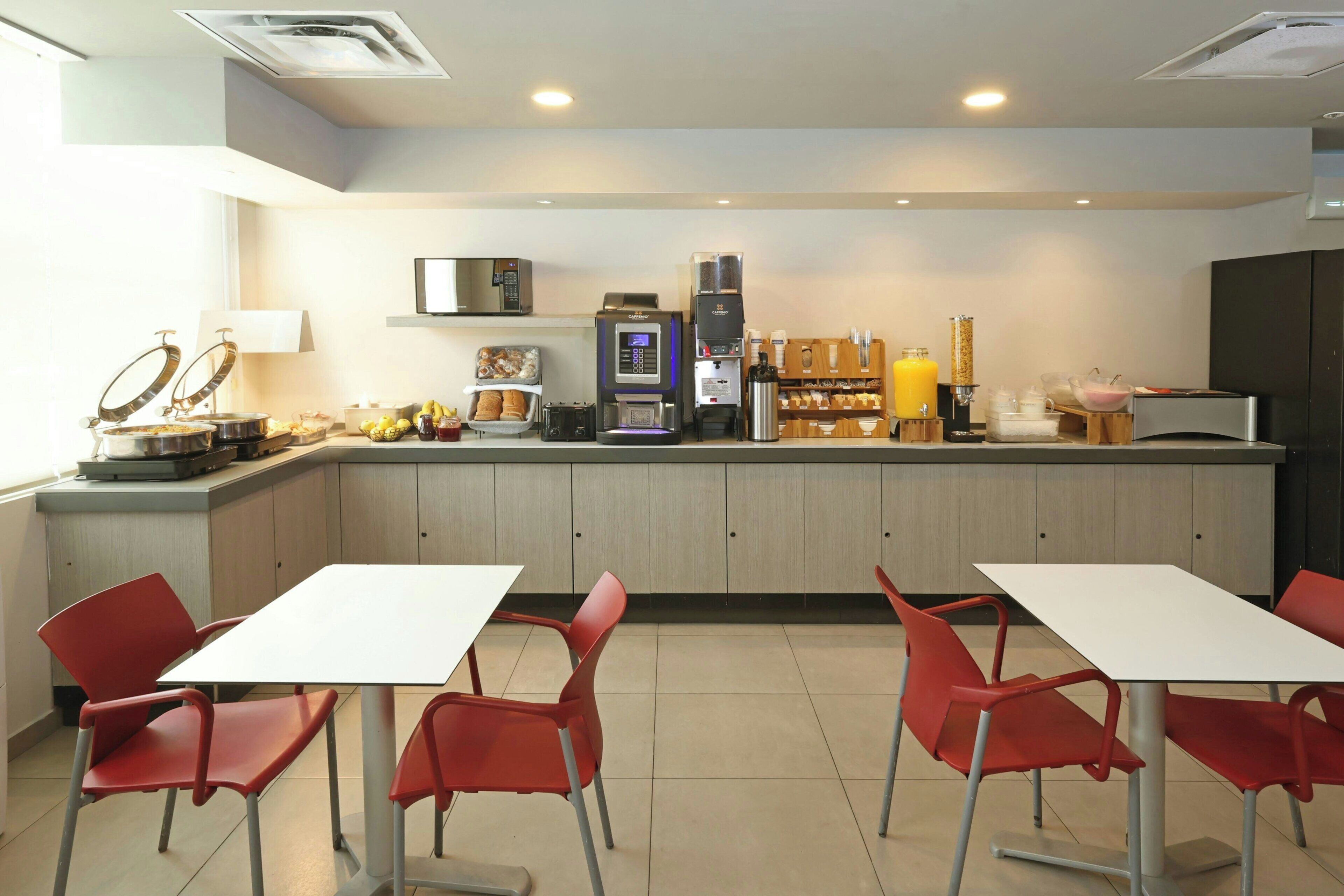 A modern kitchen with a coffee machine, microwave, and various food items on the counter. There are red chairs and a white table in the foreground.