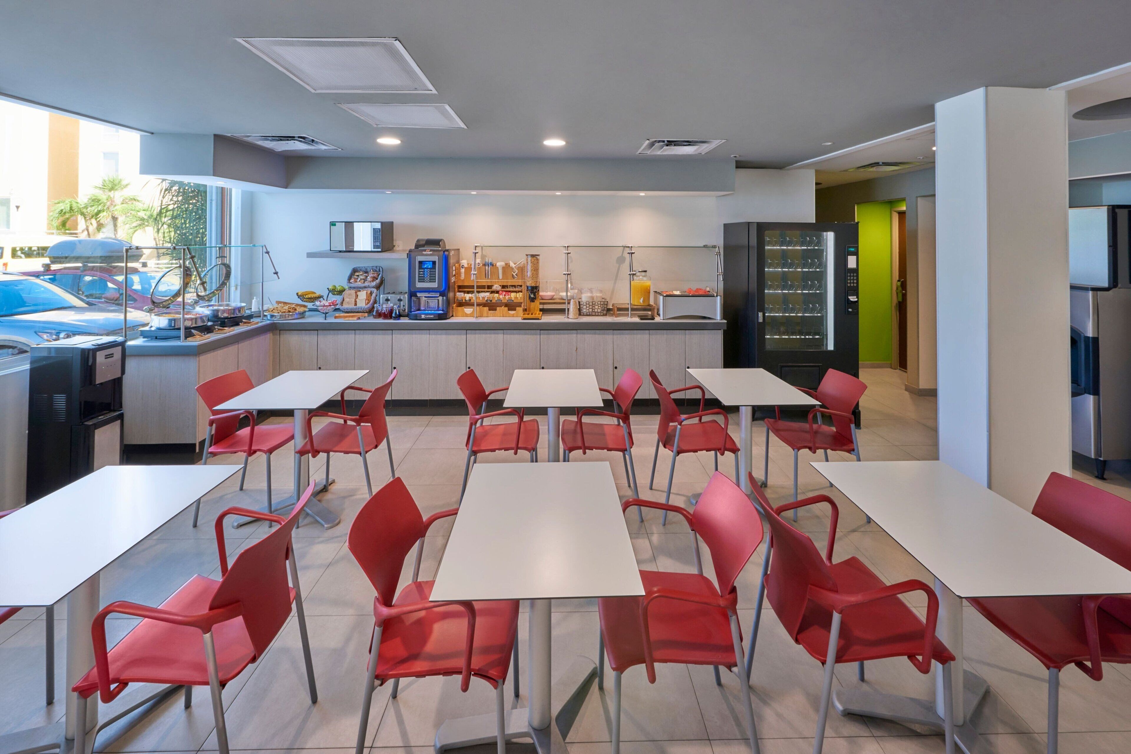Une cafétéria moderne avec des tables blanches et des chaises rouges, équipée d’une machine à café et d’un réfrigérateur.