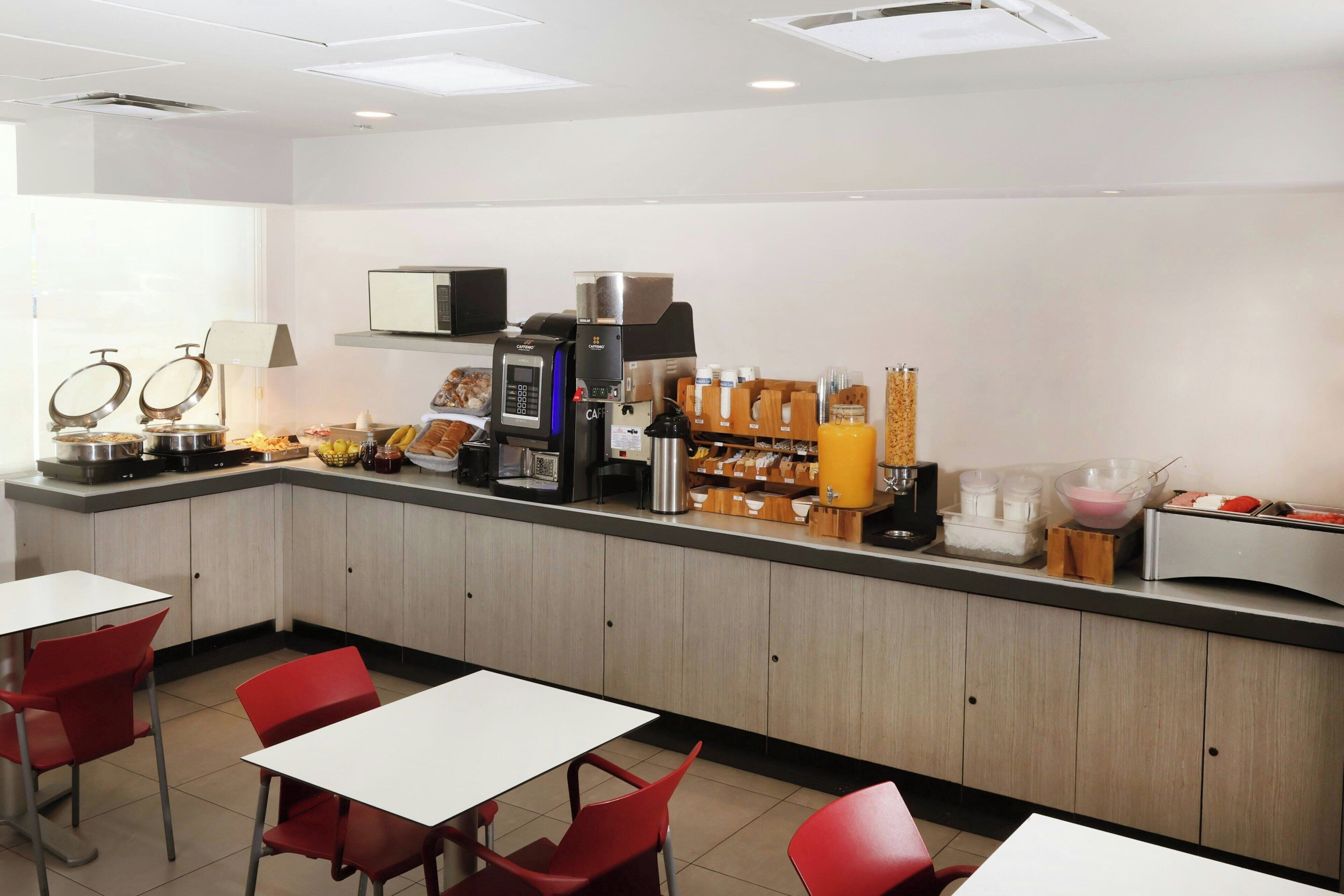 A breakfast bar with a coffee machine, fruit, and condiments. There are red chairs and a white table.