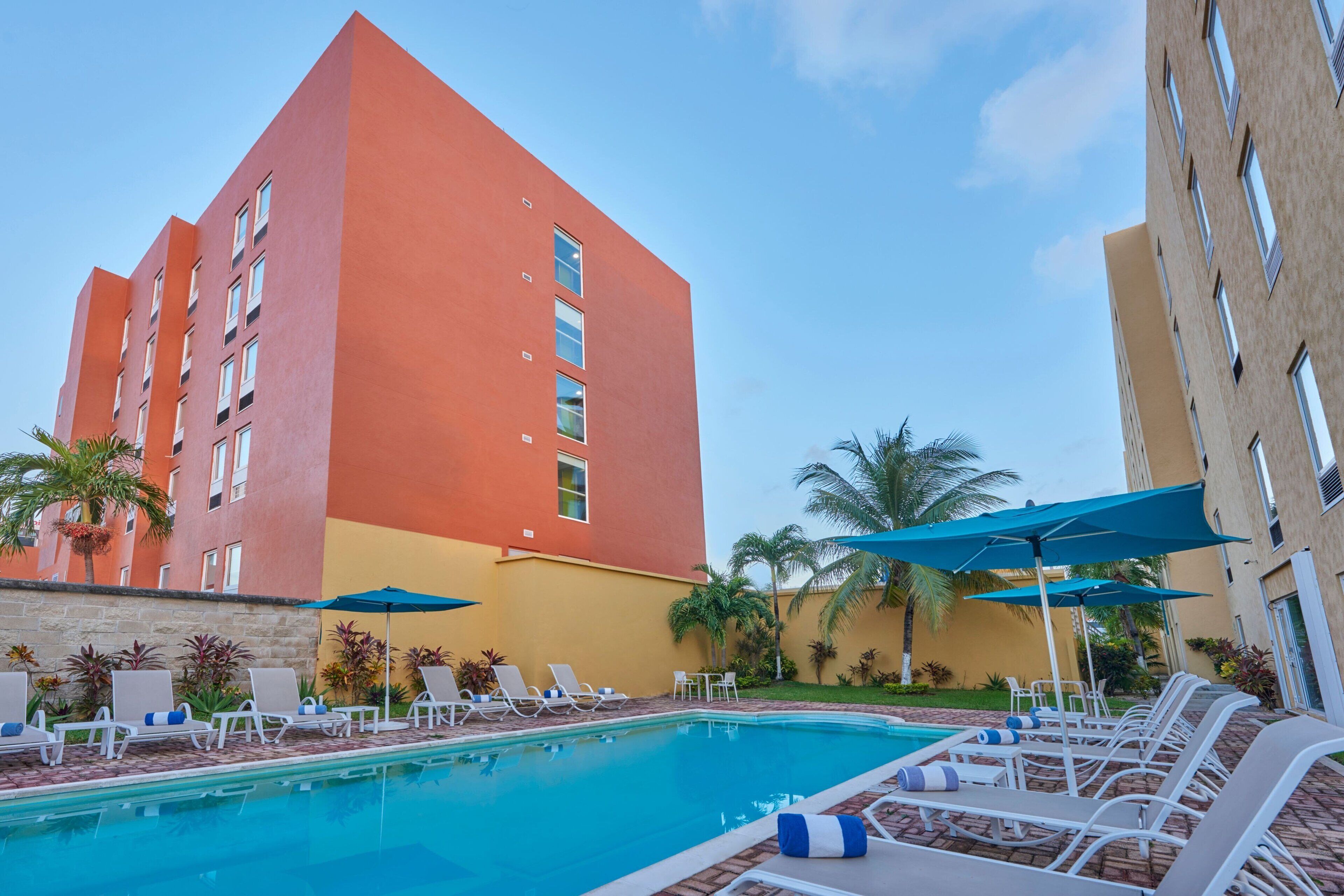 A hotel pool area with lounge chairs and umbrellas, surrounded by palm trees and a building with a red and yellow facade.
