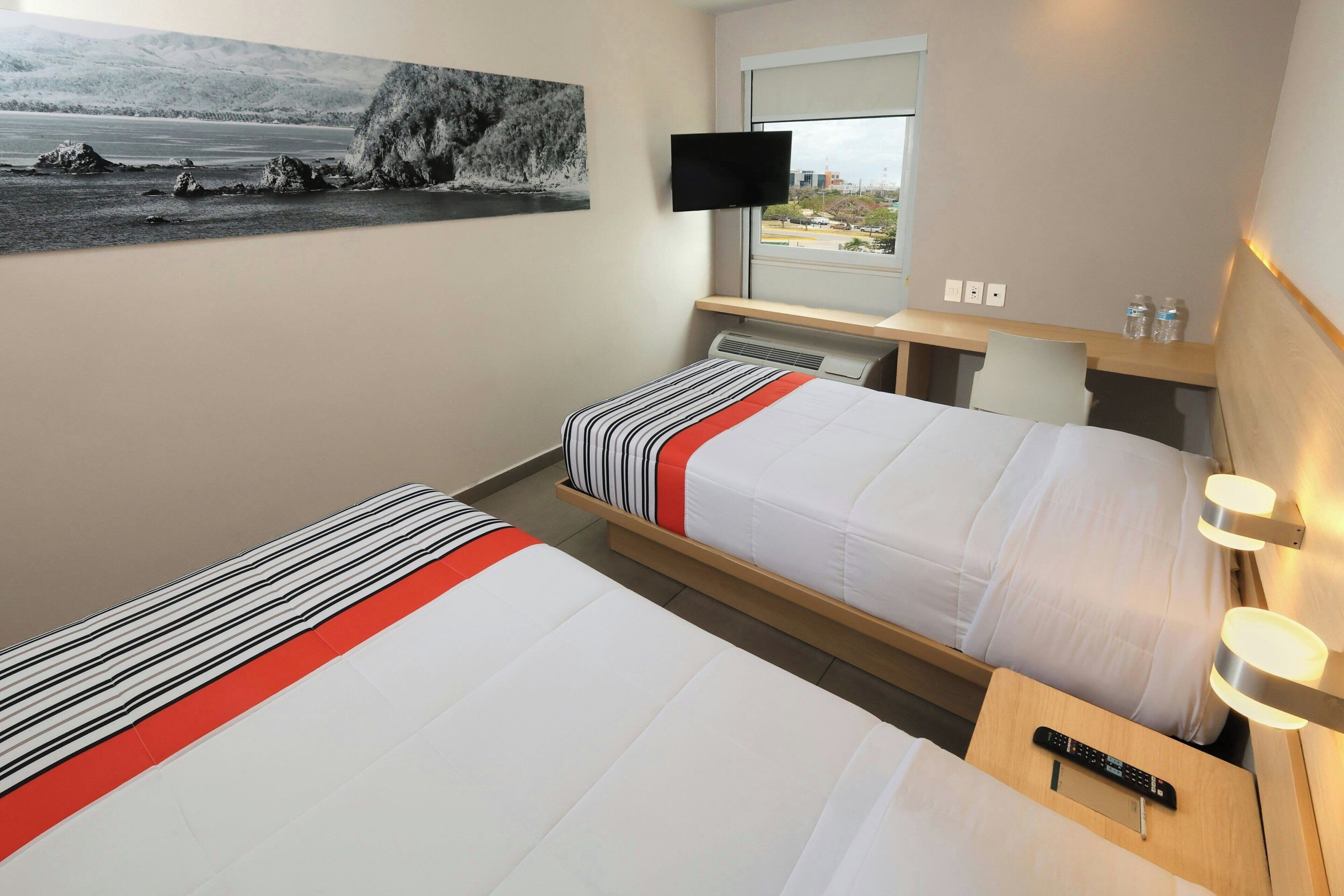 A hotel room with two beds, a TV, a window with a view, and a desk with a remote control.
