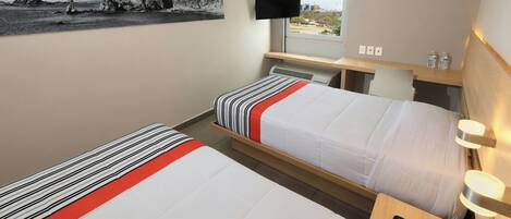 A hotel room with two beds, a TV, a window with a view, and a desk with a remote control.