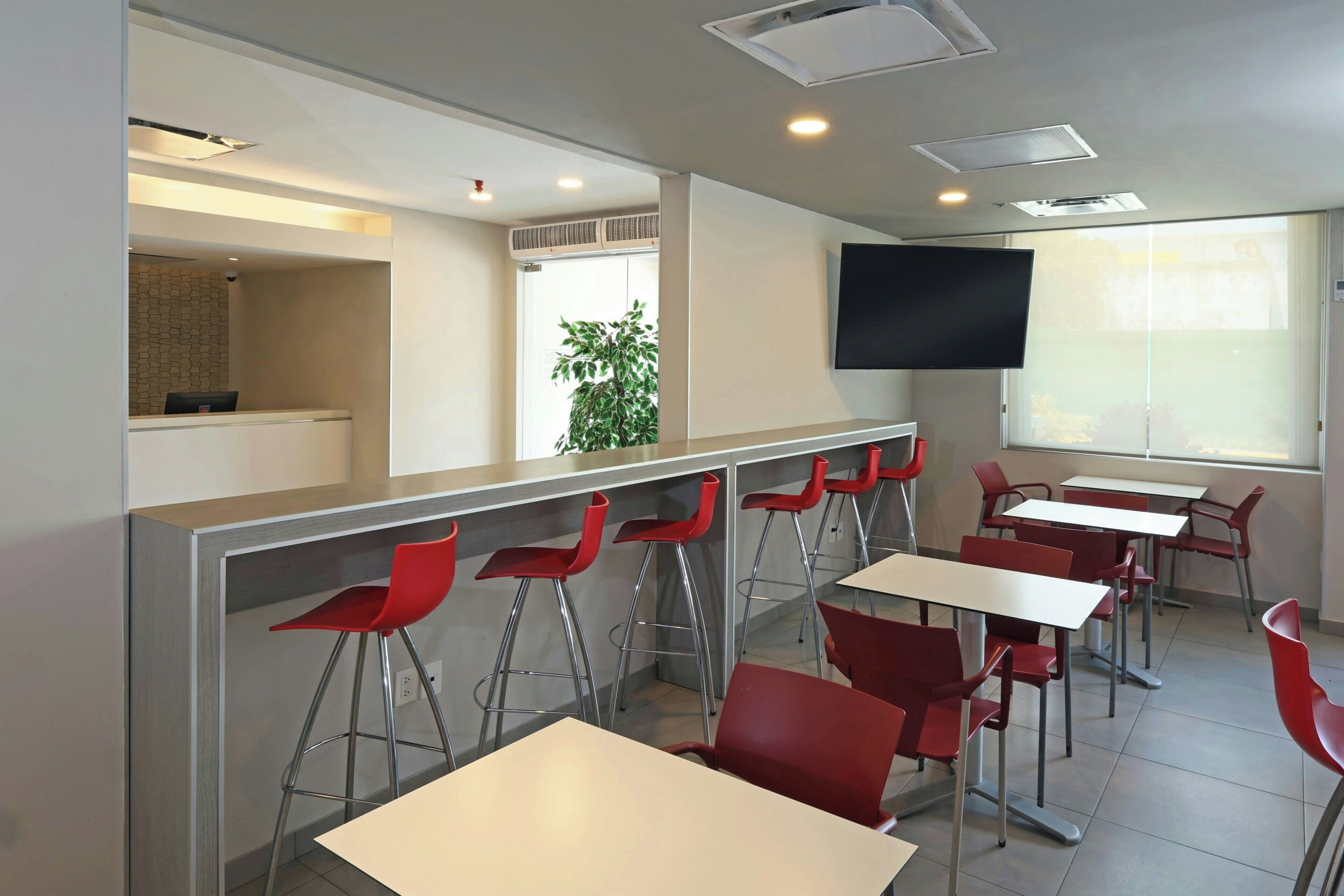 A modern dining area with high tables and red chairs, a flat-screen TV mounted on the wall, and a large window providing natural light.