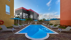 Una piscina con lettini e ombrelloni, circondata da un patio in mattoni e un edificio con tetto rosso.