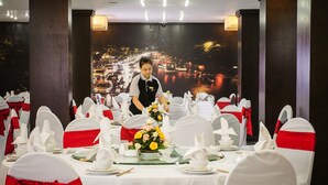 A banquet hall with round tables set for a formal event, featuring white chairs with red sashes and a backdrop of a cityscape. - Holidays-hanoi Hotel (Hanoi)