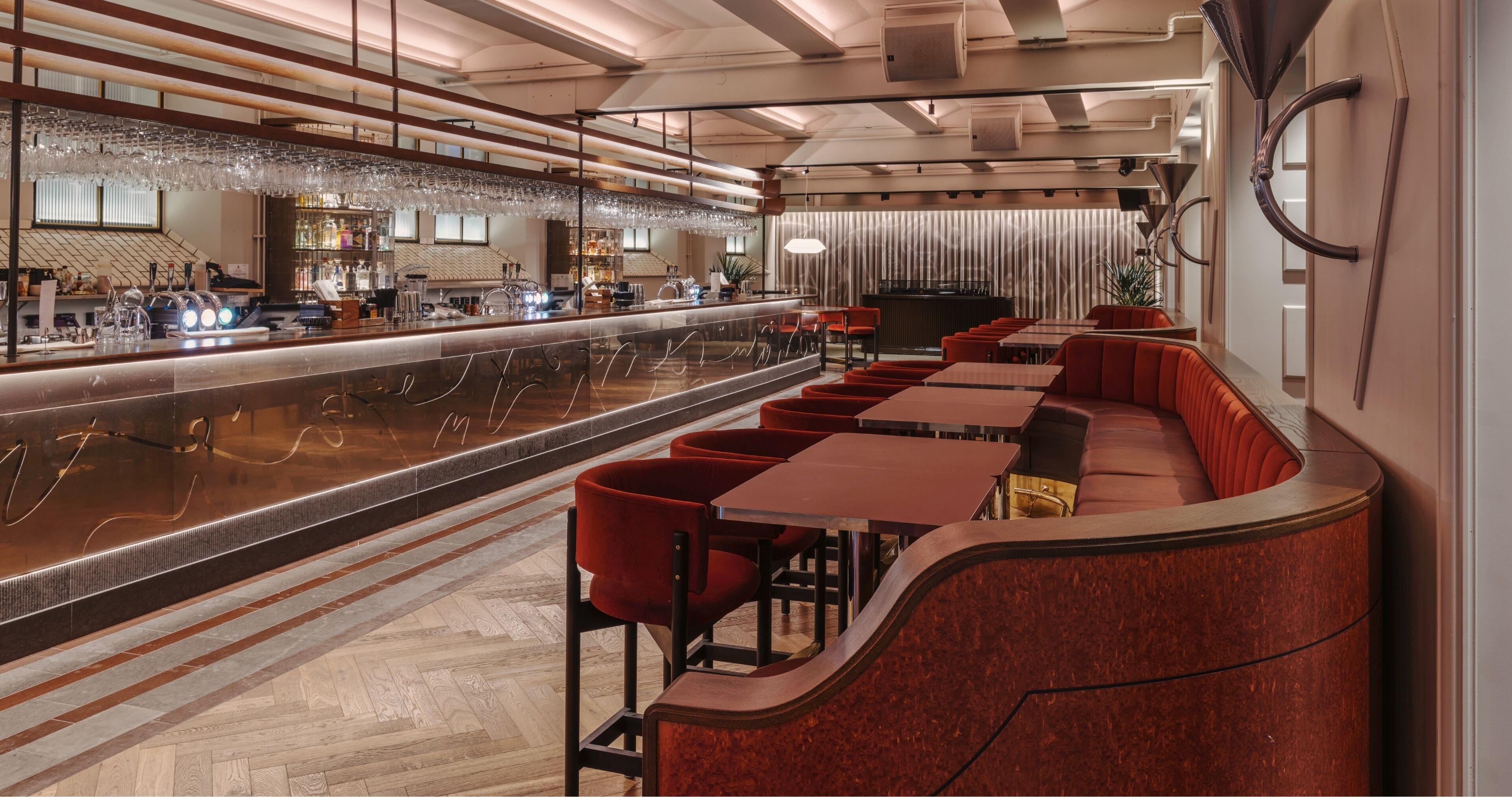 A modern bar area with a long counter, red upholstered seating, and a polished herringbone floor.