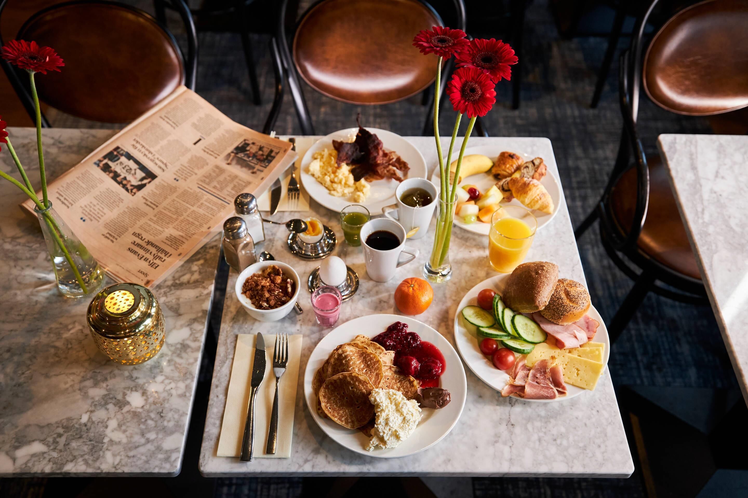A breakfast table with various dishes, including pancakes, eggs, and pastries. There are also condiments, a newspaper, and a vase with flowers.