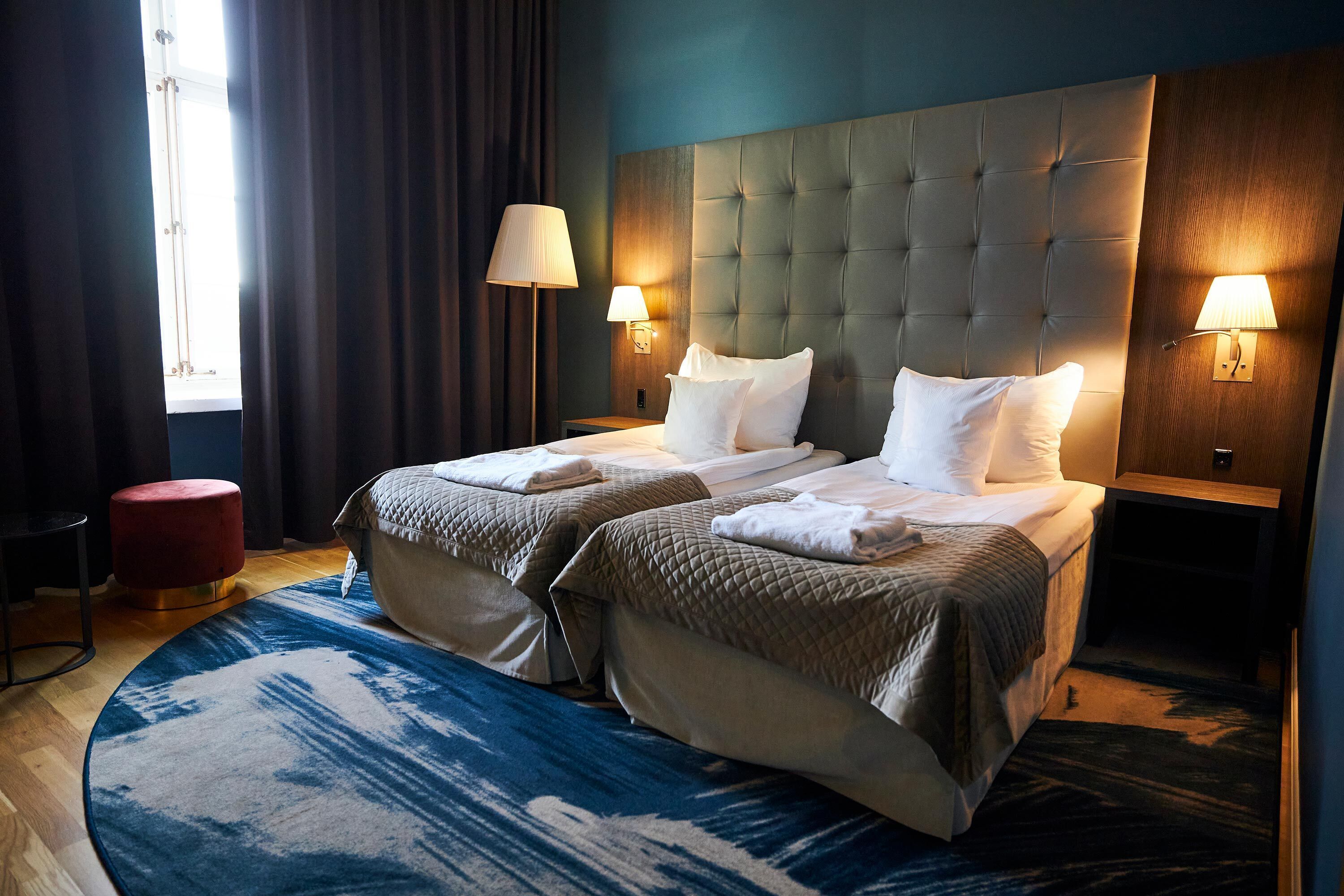 A hotel room with two beds, a red ottoman, and a large window with curtains.