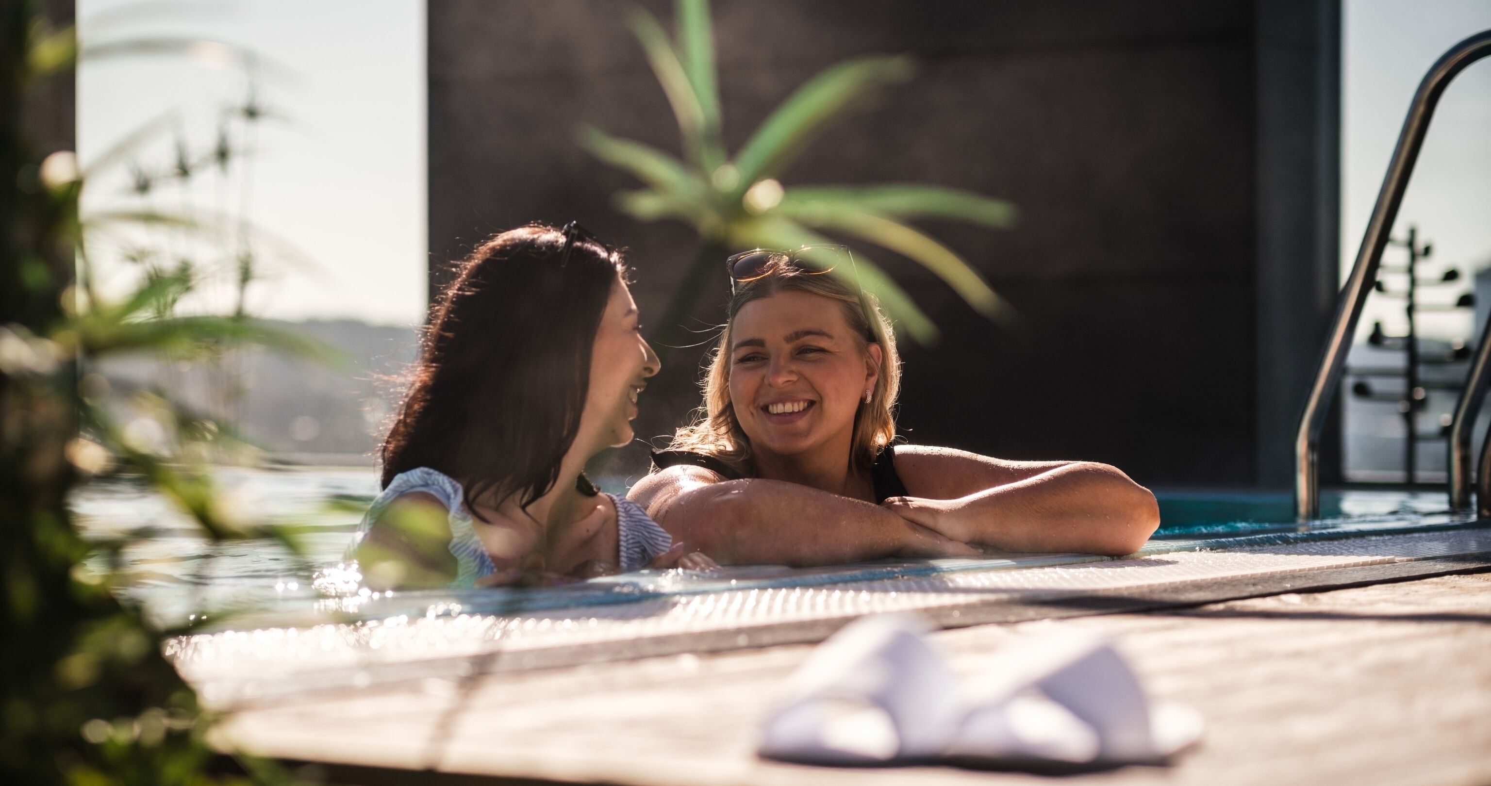 Två personer som kopplar av vid poolkanten, med en handduk och en växt i bakgrunden.