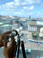 Mão segurando um telescópio, com vista para um horizonte urbano com edifícios e uma ponte.