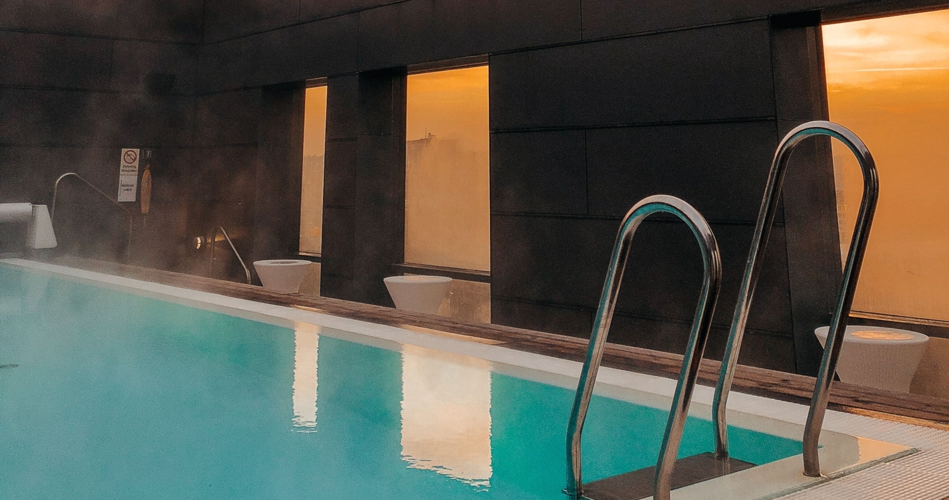 A swimming pool with metal ladders, a sauna, and a view of a sunset through the windows.
