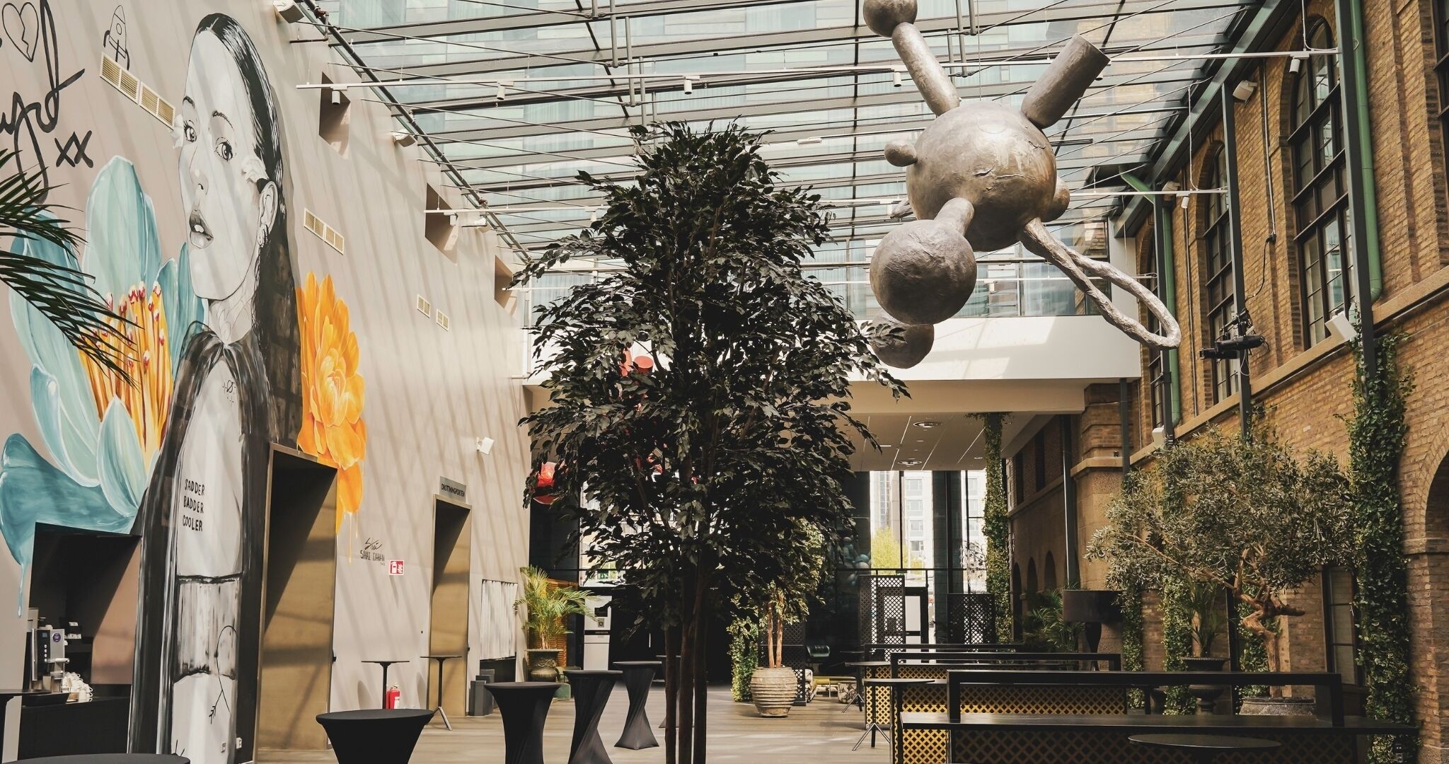 An indoor atrium with a hanging sculpture, a large tree, and a mural on the wall.