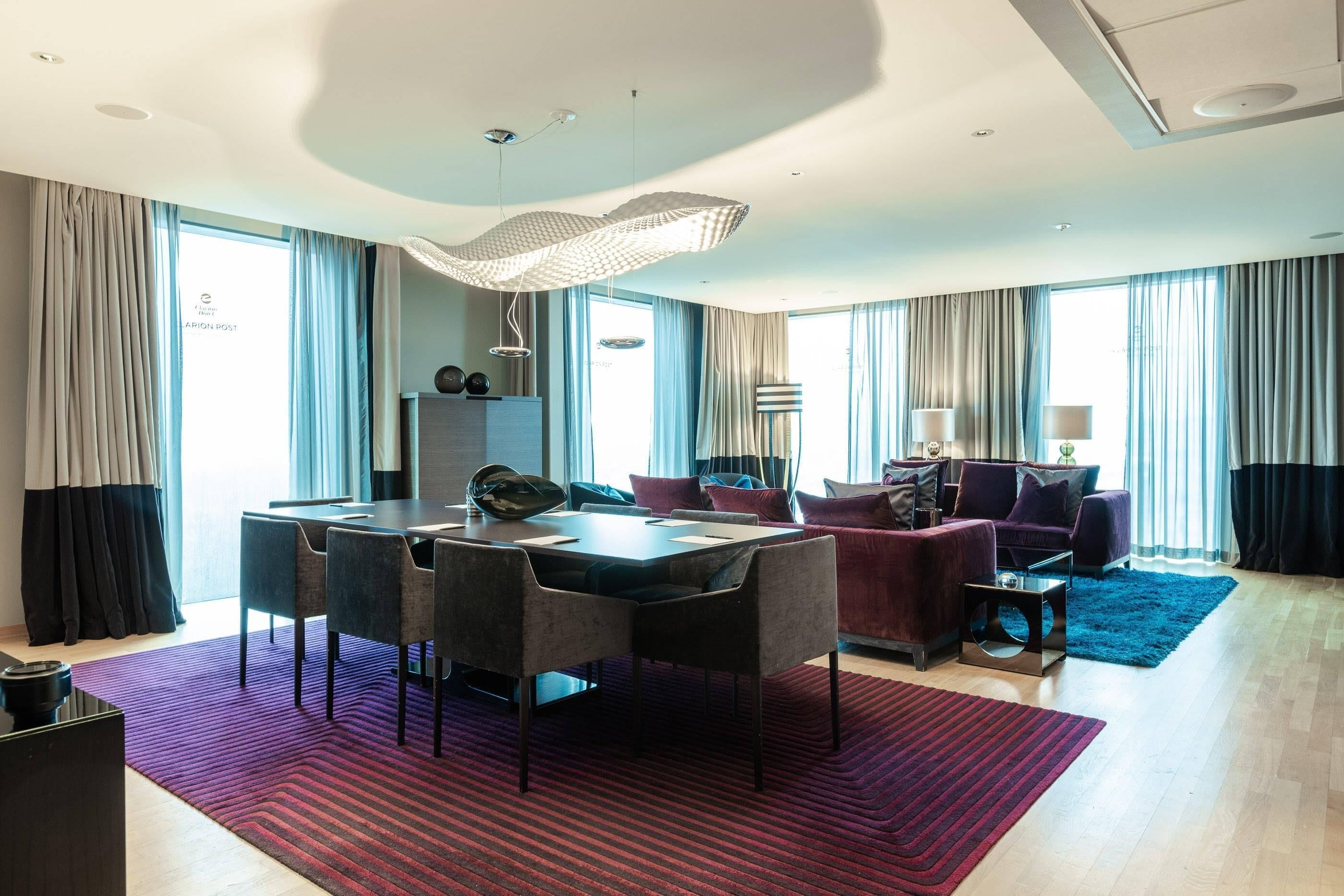 A modern hotel room with a dining area, a sofa, and a large window with curtains.